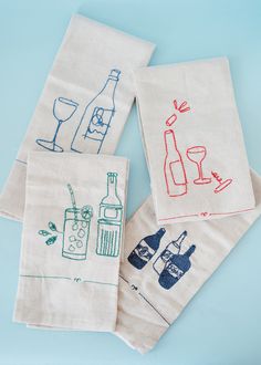 four embroidered tea towels with wine bottles and glasses on them, sitting on a blue surface