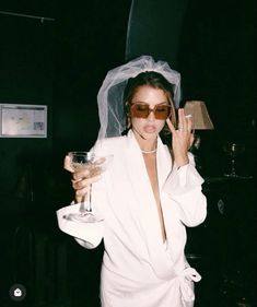 a woman in a white robe holding a wine glass and posing for the camera with a veil on her head