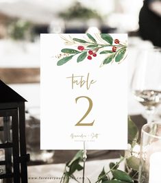 a table number is displayed on top of a place card holder with greenery and red berries