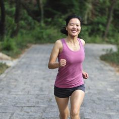 a woman running down a dirt road in the middle of the day with trees and bushes behind her