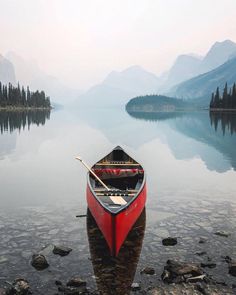 a red canoe sitting on top of a lake next to rocks and trees with mountains in the background