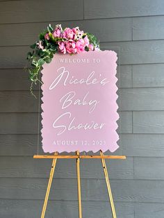 a welcome sign for a baby shower with pink flowers on it and greenery in the corner