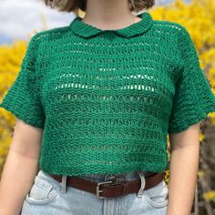 a woman standing in front of yellow flowers wearing a green knitted shirt and jeans