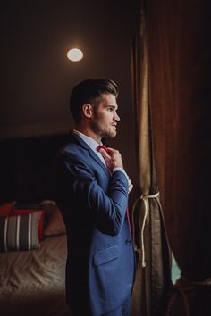 a man in a blue suit adjusting his tie while standing next to a window with curtains