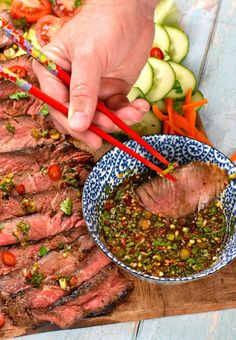 a person holding chopsticks over a bowl of food on a cutting board with vegetables and meat