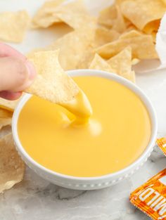 a person dipping tortilla chips into a white bowl filled with orange liquid and topped with guacamole