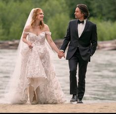 a bride and groom holding hands walking on the beach next to the water with trees in the background