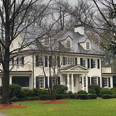 a large white house with black shutters and trees in the front yard on a cloudy day