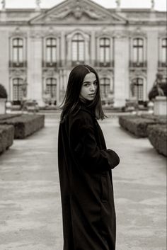 a woman standing in front of a building wearing a long black coat and boots with her hands on her hips