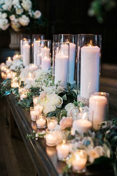 candles are lined up on a long table with white flowers and greenery around them