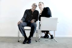 two men sitting next to each other on white chairs in front of a white wall