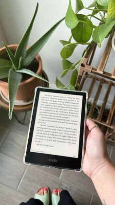 a person is holding an electronic device with a book in their hand and some potted plants behind them