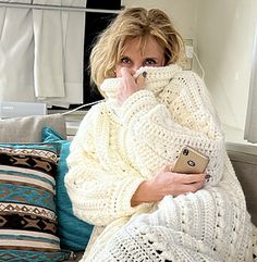 a woman sitting on a couch wrapped in a blanket and holding a remote control to her ear