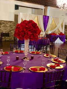 a purple table cloth with red flowers in a vase and plates on the tables set for dinner