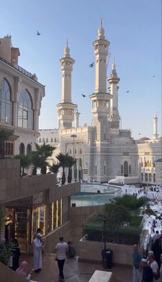 people are walking around in front of some buildings and birds flying over the building's roof tops