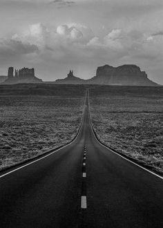 a black and white photo of an empty road in the desert with mountains in the background