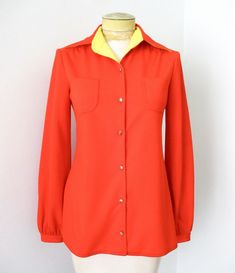 Groovy vintage 1960's or 70's tomato red textured polyester knit button front blouse from Crystal Collections. Features - navy blue stitching details - gold tone buttons - one button cuffs - chest pockets - straight hem - side splits APPROXIMATE MEASUREMENTS in inches - lying flat Size:  6 Shoulder to shoulder:  15 Sleeve, from shoulder:  23 Chest, armpit to armpit:  18 Waist:  17 Length, from shoulder:  27 CONDITION One front button is detached (it's pinned on).  Good vintage condition with lig Classic Orange Buttoned Top, Classic Orange Tops With Buttons, Retro Long Sleeve Orange Blouse, Retro Orange Long Sleeve Blouse, Red Retro Blouse, Retro Collared Blouse With Buttons, Retro Collared Button Blouse, Vintage Orange Long Sleeve Top, Vintage Orange Button-up Tops