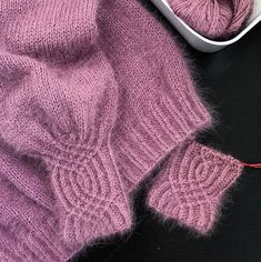 a pink sweater next to a pair of knitting needles and a ball of yarn on a black surface