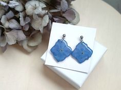 a pair of blue earrings sitting on top of a white box next to purple flowers