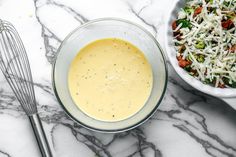 a glass bowl filled with dressing next to a metal whisk on a marble counter