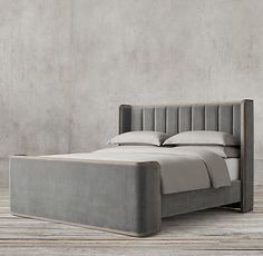 an upholstered bed with white linens and wood frame, in front of a concrete wall