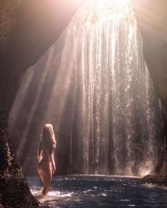 a woman standing in the water near a waterfall with sunlight streaming down on her head