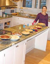 a woman standing in a kitchen with many pies on the counter and dishes on the island