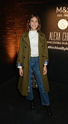 a woman standing in front of a sign wearing a trench coat and white shirt with blue jeans