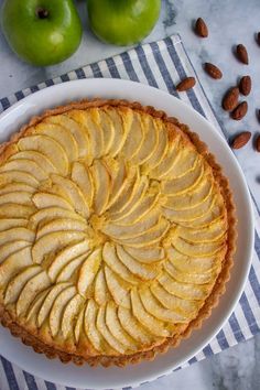 an apple pie on a white plate next to two green apples and almonds in the background