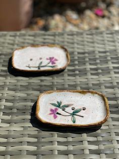 two small dishes with flowers painted on them sitting on a wicker tablecloth surface