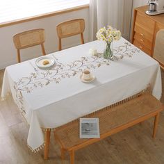 a table with a white table cloth and flowers in a vase on it, next to two wooden chairs