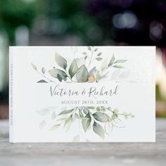 a wedding card with greenery on it sitting on top of a wooden table in front of a blurry background