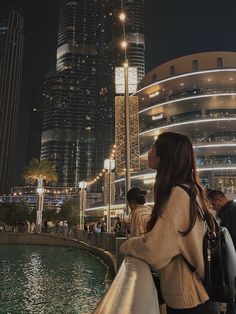 a woman standing next to a body of water in front of tall buildings at night