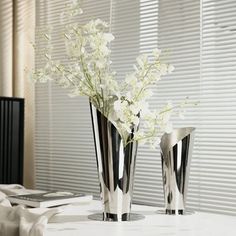 two silver vases with white flowers in them on a table next to a window