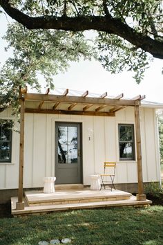 a small white house with a gray door and chair on the front porch under a large tree