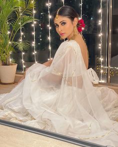 a woman sitting on top of a wooden floor wearing a white dress and gold jewelry