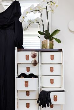 a white dresser with lots of drawers next to a flower pot and black gloves on it