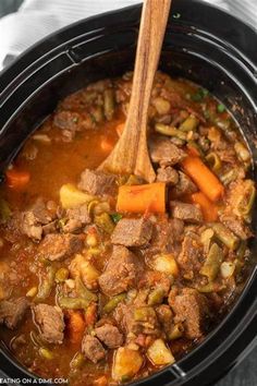 a crock pot filled with beef stew and carrots next to a wooden spoon