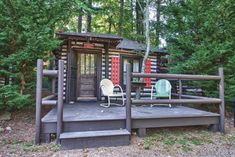 two chairs are sitting on the porch in front of a small cabin surrounded by trees