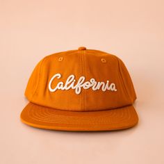 A burnt orange flat brimmed baseball hat with ivory embroidered text that reads, California across the front. Brown Summer Hats With Embroidered Logo, Brown Hats With Embroidered Logo For Summer, Brown Hat With Embroidered Logo For Summer, Brown Dad Hat With Letter Print, Brown Dad Cap With Letter Print, Vintage Cotton Hats For Travel, Vintage Cotton Travel Hat, Vintage Brown Hat For Everyday, Everyday Vintage Brown Hat