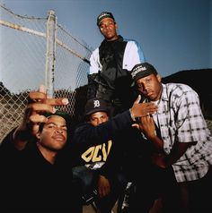 four young men posing for a photo in front of a chain link fence with one pointing at the camera