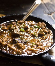 a skillet filled with meat and vegetables on top of a wooden table next to utensils