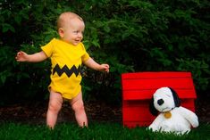 a baby standing in the grass next to a stuffed dog and a red box with a black and yellow design on it