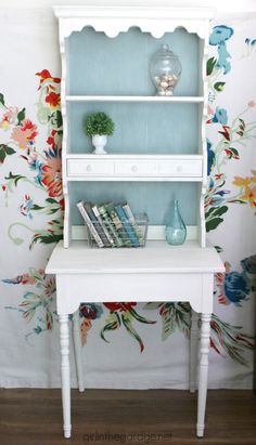 a white book shelf with books and vases on it in front of a floral wallpaper