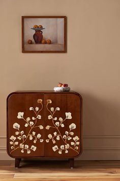 a cabinet with flowers painted on it in front of a painting hanging on the wall