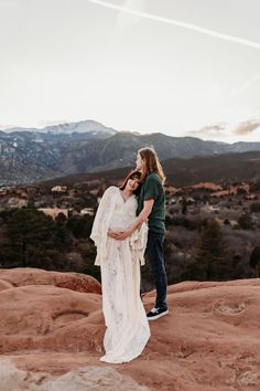 a pregnant woman standing next to a man on top of a mountain with mountains in the background