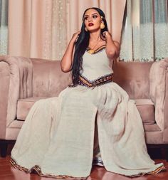 a woman sitting on top of a couch wearing a white dress and gold jewelry with her hands behind her head