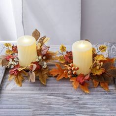 two candles with autumn leaves and berries are on a wooden table next to each other