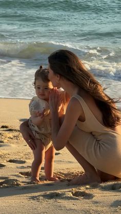 a woman kneeling down next to a baby on the beach