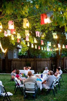 a group of people sitting around a table in the grass with lights strung above them